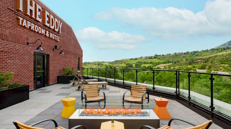 The image shows an outdoor patio at "The Eddy Taproom & Hotel" with lounge chairs, yellow and orange tables, and a rectangular firepit, surrounded by greenery.