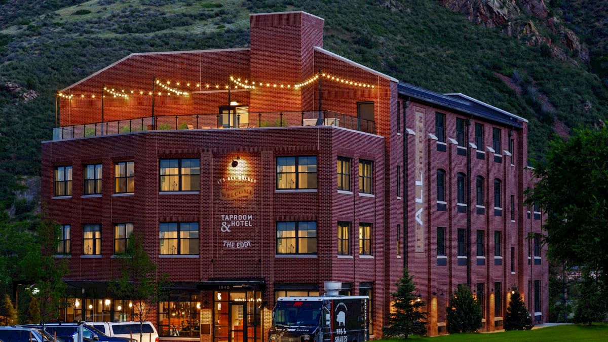 A multi-story brick building, likely a hotel or inn, with string lights on the rooftop patio and surrounded by lush green hills in the background.