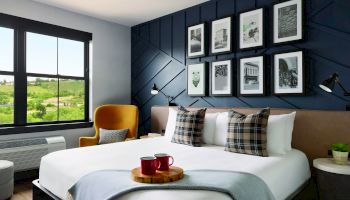 A modern bedroom with a king-size bed, decorative pillows, framed pictures on a dark accent wall, a yellow chair, and two mugs on a tray on the bed.