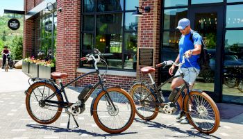 A person in a blue shirt and white cap stands beside two parked bicycles in front of a brick building with large windows and plants outside.