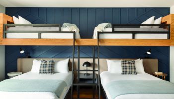 A modern hotel room with two full beds on the lower level and two twin beds on a mezzanine, dark blue paneled wall, and wooden accents.