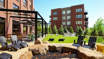 An outdoor seating area with chairs around a fire pit, greenery, and a game area in front of brick buildings on a sunny day.