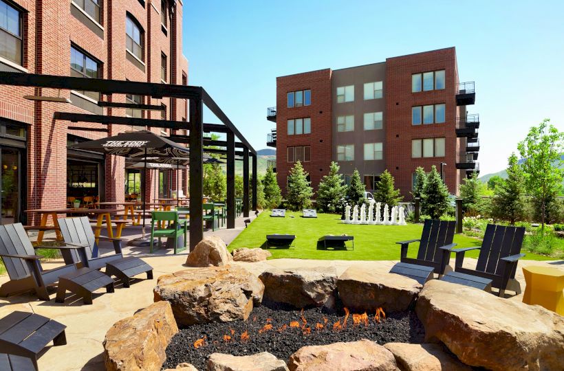 An outdoor seating area with chairs around a fire pit, greenery, and a game area in front of brick buildings on a sunny day.