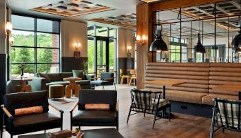 The image showcases a modern, elegantly furnished lounge area with large windows, comfortable seating, and a blend of wooden and industrial elements.