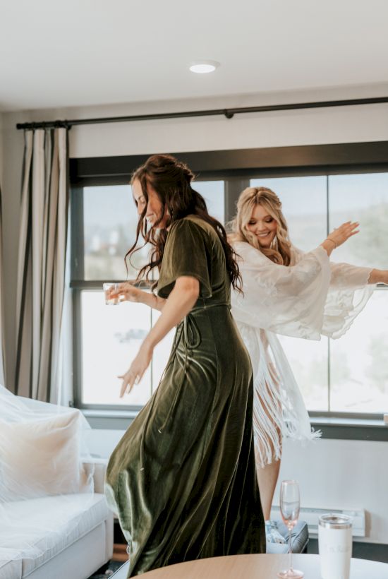 Two women in elegant dresses laugh and celebrate indoors, arms out as they enjoy champagne at a bright event table.