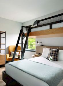 The image shows a modern bedroom featuring a double bed, a lofted bed with a ladder, a wardrobe with robes, and a cozy chair by the window.