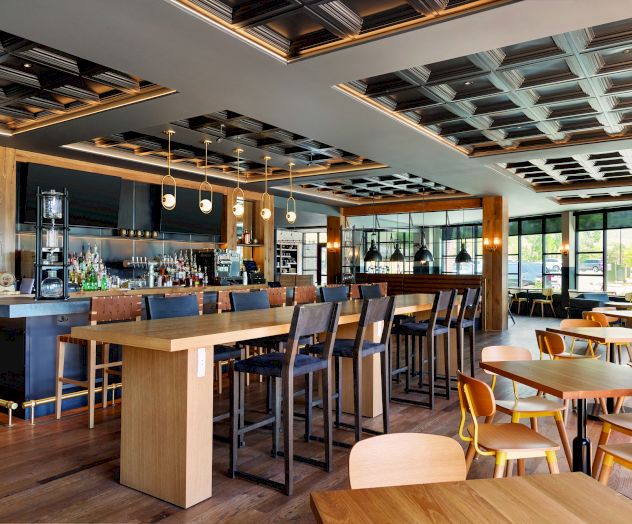 The image shows a modern, upscale restaurant interior with wooden tables and chairs, a bar counter with stools, and intricately designed ceiling panels.